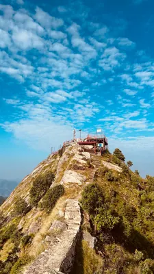 A narrow rocky path climbs a steep ridge toward a small hilltop temple beneath blue sky.
