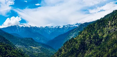 Green valley slopes and small hillside settlements beneath snowy mountains, white clouds and vivid blue sky.