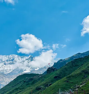 White clouds drift above snowy peaks and a green mountainside beneath a broad blue sky.