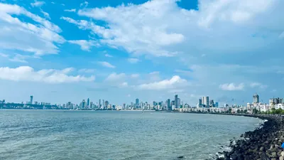 A coastal city skyline curves around blue water under a bright sky filled with scattered clouds.