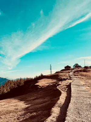 A pale path winds up a warm, dry hill toward a small building beneath sweeping white clouds.